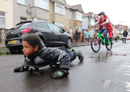 Boy lying on skateboard in street with child cyclist behind